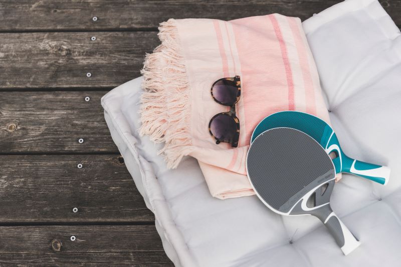 Detailaufnahme von zwei Outdoor-Tischtennis-Schlägern, die auf einem Outdoor-Kissen und einem Badetuch liegen. Eine Sonnenbrille liegt ebenfalls auf dem Badetuch.