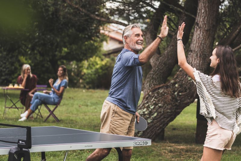 Ein älterer Herr steht mit einer Jugendlichen an der Tischtennisplatte auf einer Seite. Sie geben sich ein High-Five und freuen sich. Im Hintergrund ist ein großer Baum zu sehen, sowie ein kleiner Beistelltisch, an dem zwei Frauen sitzen, die sich unterhalten und zu den beiden an der Tischtennisplatte rüber sehen.