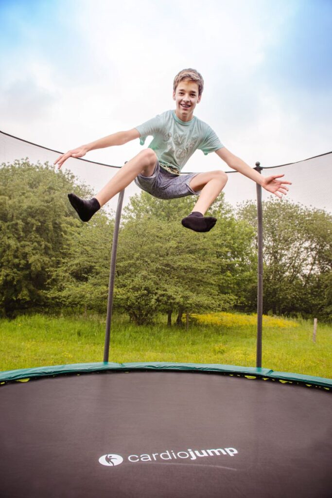 Ein Junge in kurzer Hose, T-Shirt und Socken springt in die Luft auf einem cardiojump-Trampolin, welches in einerm Garten steht. Im Hintergrund sind Bäume zu sehen. der Himmel ist leicht bewölkt.