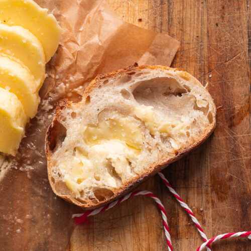 Une tranche de pain rustique beurrée repose sur une planche en bois, à côté de plusieurs morceaux de beurre et d'un bout de ficelle rouge et blanche.