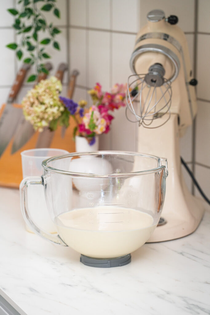 A glass mixing bowl containing a liquid batter is placed on a marble countertop, next to a stand mixer, with flowers and knives in the background.