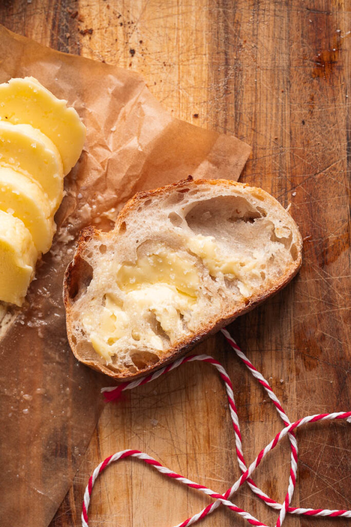 A slice of buttered rustic bread rests on a wooden board, next to several pieces of butter and a piece of red-and-white string.