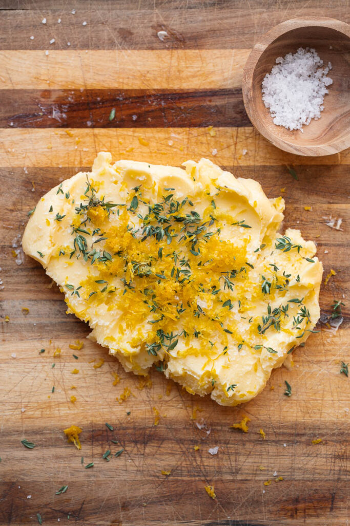 A pat of butter sprinkled with lemon zest and fresh thyme rests on a wooden cutting board, next to a small wooden bowl containing coarse salt.