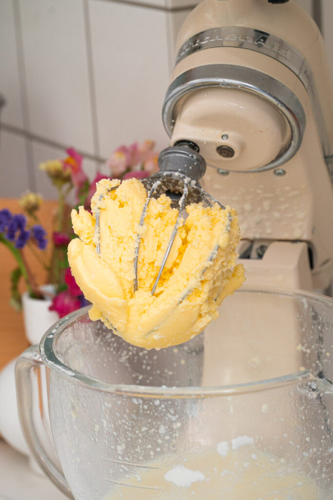 A stand mixer fitted with a whisk is holding freshly churned butter over a glass mixing bowl. Flowers can be seen in the background.