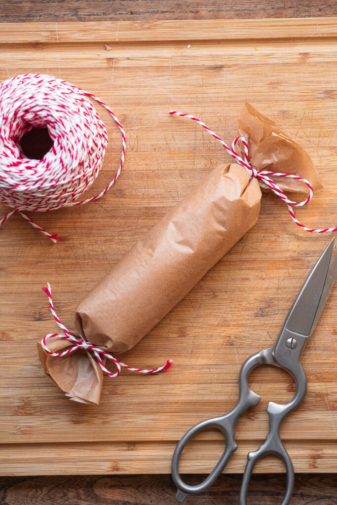 A cylindrical package wrapped in kraft paper and tied with red and white string rests on a wooden surface, next to a pair of scissors and a spool of string.