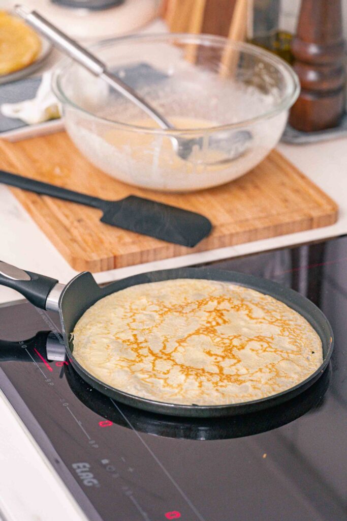A crêpe cooking in a skillet on a stove, with a glass mixing bowl containing crêpe batter, a spatula, and some extra batter in the background.