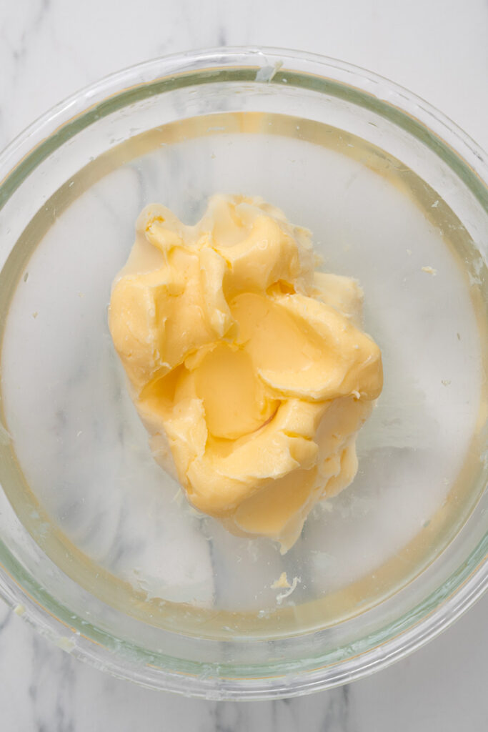 A glass bowl containing a mound of pale yellow butter, sitting on a white marble countertop.